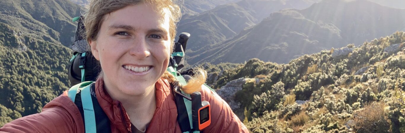 A smiling woman with a hiking backpack taking a selfie with a mountain backdrop.