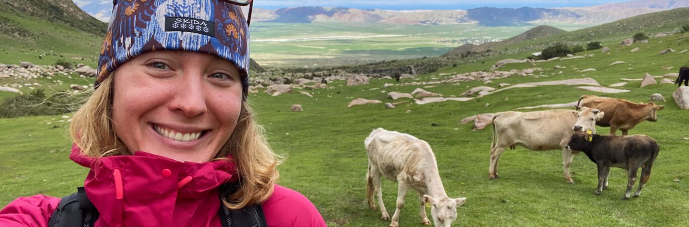 Taylor Nydam taking a selfie with a large green field and cows in the background in Kyrgyzstan.